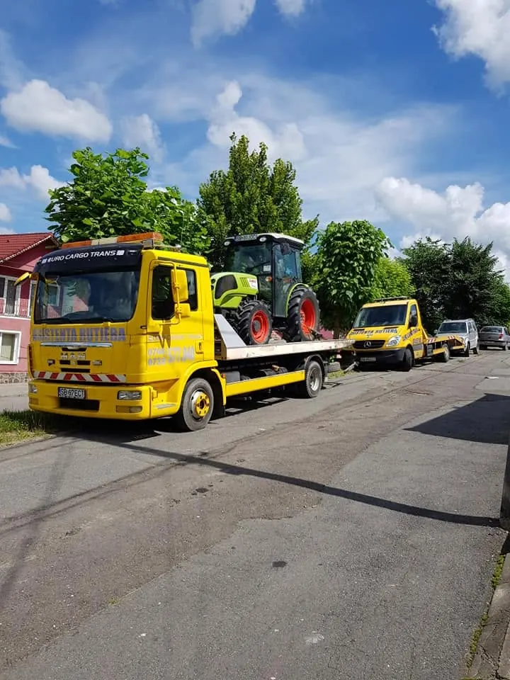 Tractor încărcat pe platformă, lângă un al doilea camion galben